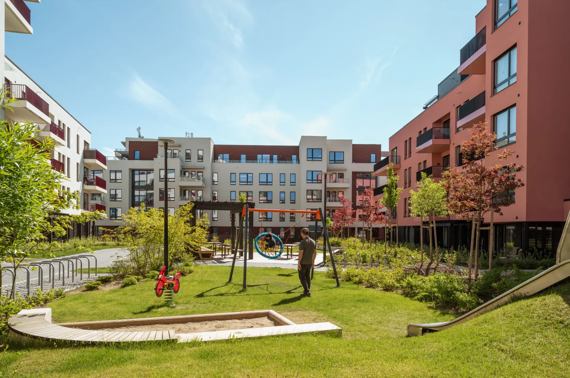 Gårdskvartalet med barnehage på Skårer skårerbyen. fasadene har hvit og rød/ rosa puss, farget balkongrekkverk, og farget beslag på dekkeforkanter, slik at balkongene i større grad er en del av fasadelivet, og gir et skulpturelt uttrykk. Utearealet er grønt og frodig.