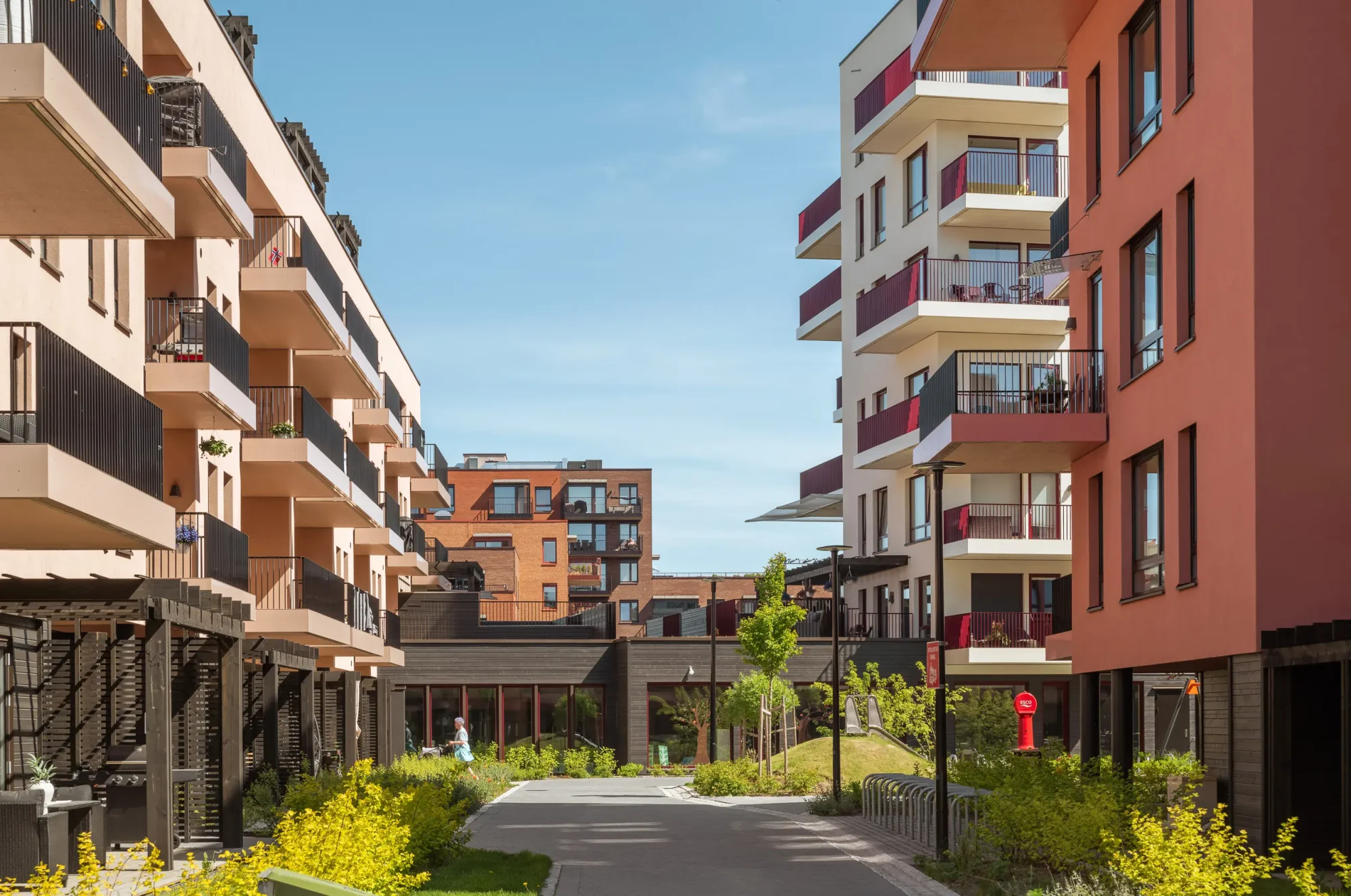 Gårdskvartalet med barnehage på Skårer skårerbyen. fasadene har hvit og rød/ rosa puss, farget balkongrekkverk, og farget beslag på dekkeforkanter, slik at balkongene i større grad er en del av fasadelivet, og gir et skulpturelt uttrykk. Utearealet er grønt og frodig.